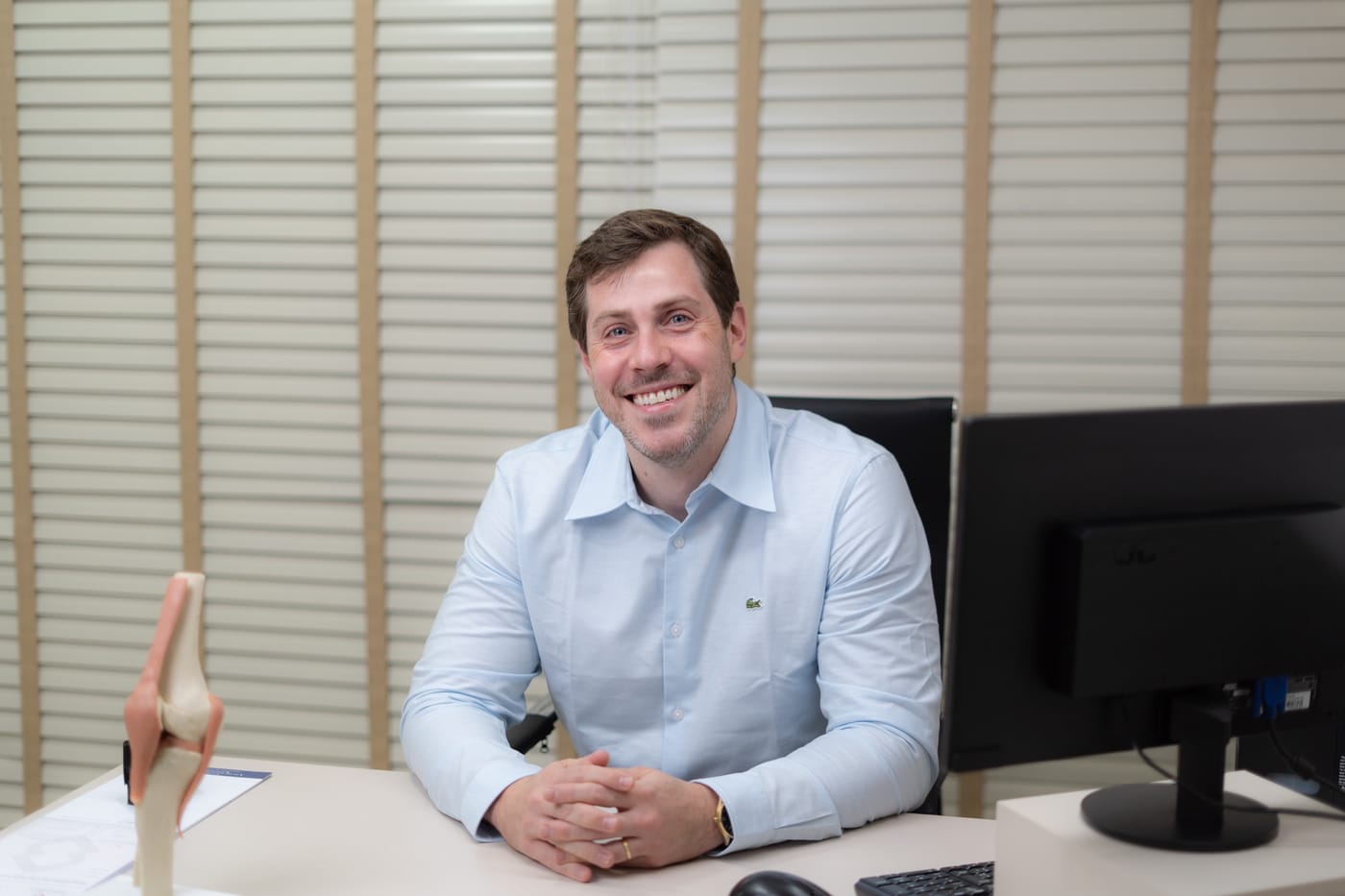 Dr. Fernando Hehn sorrindo à mesa do consultório, com um modelo anatômico do joelho ao lado